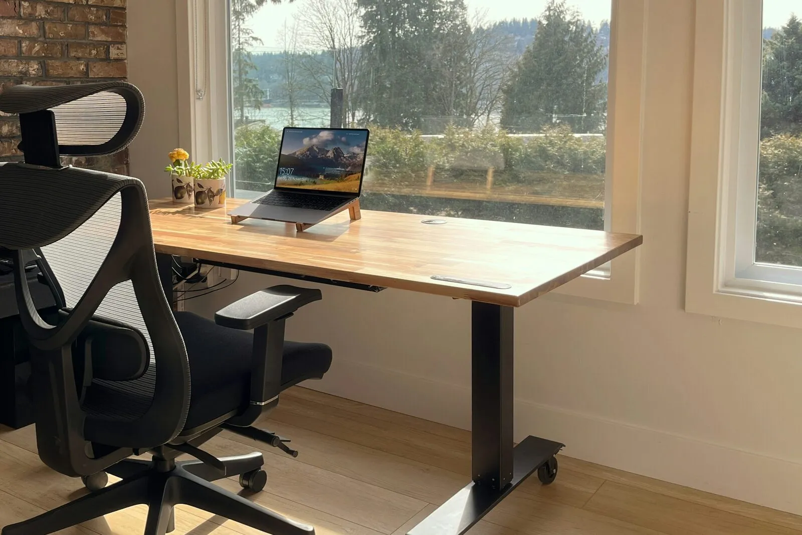 A laptop on a wooden desk by a sunny window, with trees and water outside