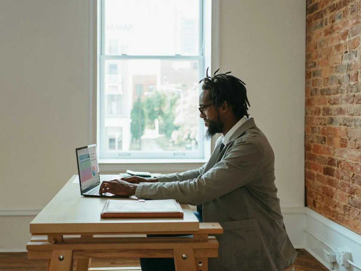 A man at a wooden desk typing a spreadsheet on his laptop, a folder and calculator beside him
