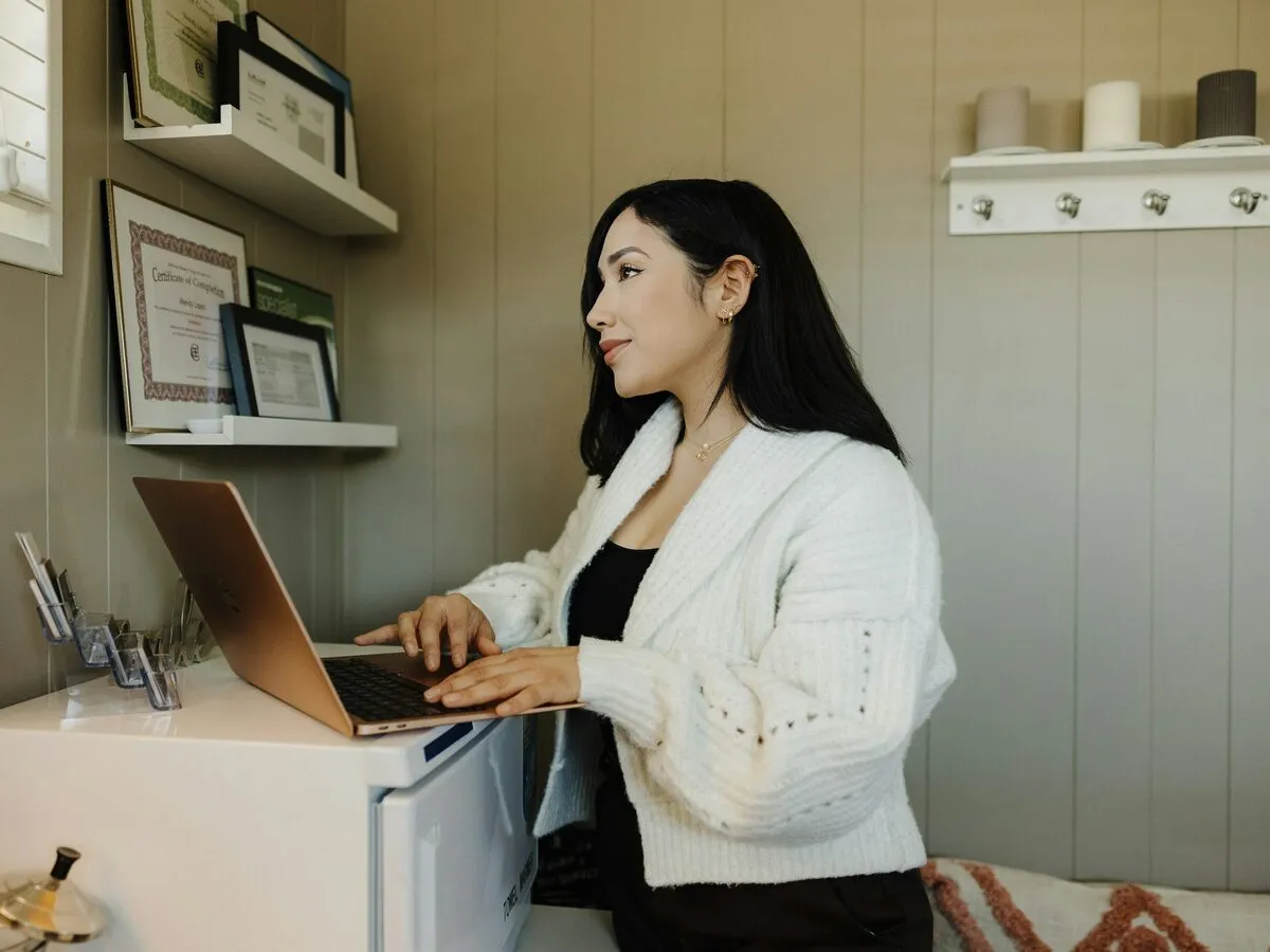 A business owner in a white cardigan typing on a laptop at her studio counter