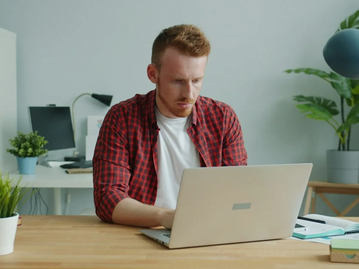 A man in a flannel shirt working on a laptop at his desk